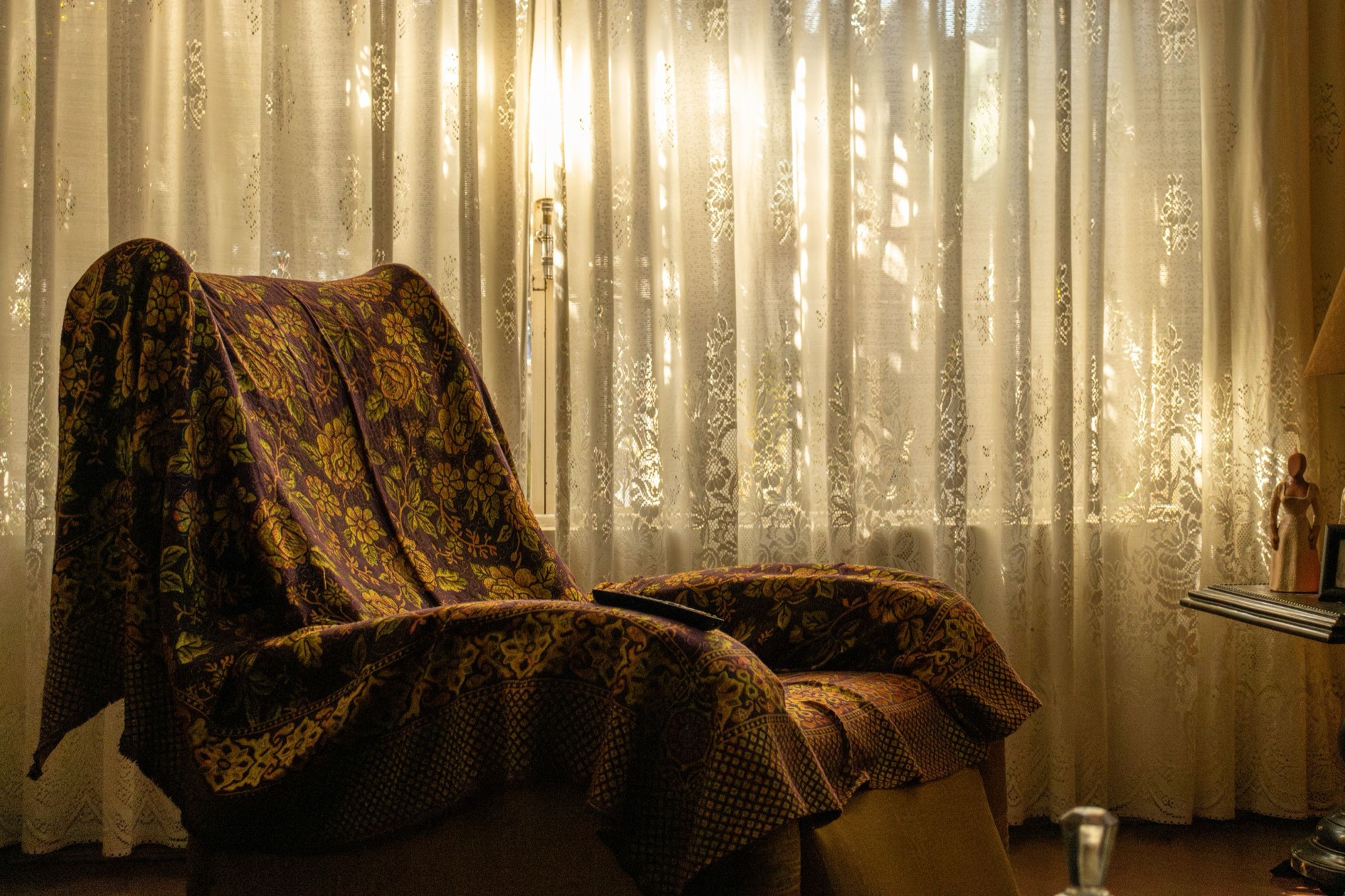 An armchair draped with a floral throw beside a lace-curtained window, warm afternoon light filling the room.